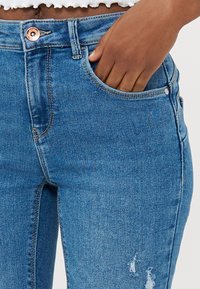 Jeans de mezclilla azul ajustados con cintura alta, que presentan un botón de cobre, bolsillos frontales y un ligero desgaste cerca del muslo.