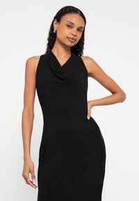 Γυναίκα με σγουρά μαλλιά που φοράει μαύρο αμάνικο φόρεμα με ντραπέ λαιμόκοψη, ποζάρει με το ένα χέρι στη μέση της μπροστά σε απλό φόντο.