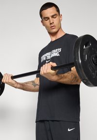 T-shirt sportivo nero con testo bianco, vestibilità standard. Persona solleva un bilanciere nero. Tatuaggi notevoli visibili sull'avambraccio. Pantaloni corti scuri.