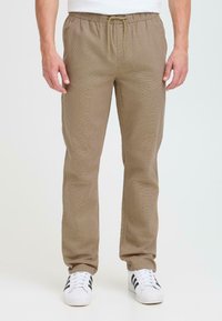 Brown, textured, elastic waistband trousers with a drawstring and side pockets, paired with white sneakers featuring black stripes.