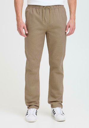 Pantalones de cintura elástica de color marrón, texturizados, con cordón y bolsillos laterales, combinados con zapatillas blancas con rayas negras.