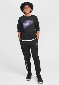 Sudadera negra de Nike con logo azul-naranja y el texto "EST. 1972", combinada con pantalones cargo negros que tienen bolsillos laterales y acentos de texto en blanco.