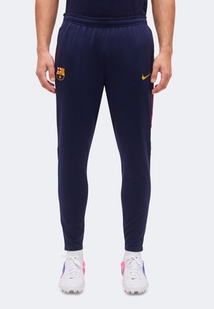Homme portant un pantalon de sport slim bleu marine avec le blason du FC Barcelone sur la cuisse gauche et le logo jaune Nike sur la cuisse droite, chaussures à crampons blanches.