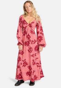 Vrouw met lang blond haar, gekleed in een roze en rode bloemenmaxi-jurk met lange, opgeblazen mouwen en zwarte schoenen, staande tegen een effen achtergrond.