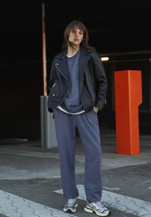 Zwarte leren motorjack over een grijze sweatshirt, gecombineerd met grijze joggingbroek en grijze sneakers. Achtergrond heeft een oranje geometrische structuur.