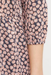 Zbliżenie na materiał z białym i pomarańczowym wzorem kwiatowym na granatowym tle, ukazujące zebrany szew oraz krótką rękaw na ramieniu osoby.