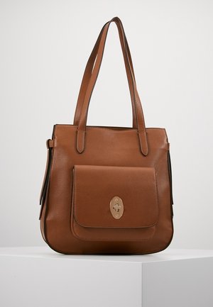 Sac à bandoulière en cuir marron avec poche avant à rabat et fermoir tournant doré, posé sur un socle blanc.
