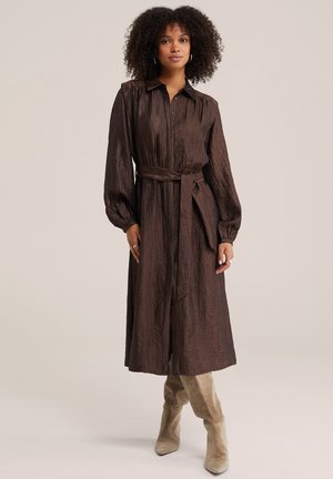 Femme aux cheveux bouclés portant une longue robe marron boutonnée avec une ceinture à la taille et des bottes en daim beige, debout devant un fond uni.