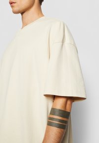 Beige bomulls t-skjorte med avslappet passform og senket skuldre. Har en subtil brodert logo på brystet og en jevn tekstur.