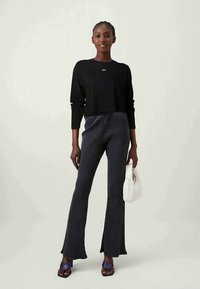 Μαύρο κοντό μακρυμάνικο μπλουζάκι με λογότυπο μπροστά, συνδυασμένο με σκούρες ριγέ φαρδιές παντελόνες. Το μοντέλο κρατάει μία λευκή τσάντα και φοράει μωβ σανδάλια.