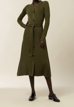 Persona de piel oscura modelando un vestido de punto acanalado verde oliva de manga larga con botones, cinturón y zapatos de charol negro.