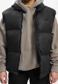 Czarna kamizelka puchowa z zamek błyskawicznym z przodu i wysokim kołnierzem, noszona na szarym swetrze. Posiada pikowane kieszenie do przechowywania i subtelne logo.