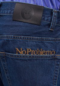 Σκούρο μπλε τζιν με καφέ κεντημένο «No Problemo» στην πίσω τσέπη και μαύρο δερμάτινο πατς με κυκλικό λογότυπο πάνω από τη μέση.
