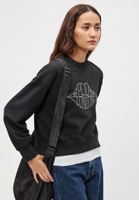 Μαύρο φούτερ με γραφικό σχέδιο χειλιών και κείμενο, συνδυασμένο με μπλε τζιν και σκούρο τσάντα ώμου. Μαλακό ύφασμα με άνετη εφαρμογή.