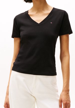 Kobieta ubrana w dopasowaną czarną koszulkę z krótkim rękawem i dekoltem w kształcie litery V, z małym logo na lewej piersi, oraz białe spodnie.