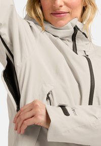 Veste imperméable gris clair avec un col haut, zips noirs, poignets réglables et poches latérales, présentant un design épuré et minimaliste.