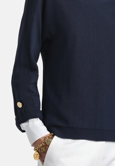 Pulover din lână bleumarin cu design texturat, mâneci rulate și un accent cu nasture auriu. Purtat cu o cămașă albă și o brățară cu monedă aurie.