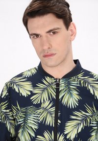 Giacca zip-up blu navy con stampa di foglie di palma in verde. Include un colletto nero e un cursore rotondo per la chiusura.