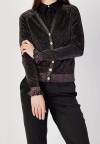Cardigan noir avec paillettes, présentant un col en V, cinq boutons et des poignets volants. Associé à un pantalon noir bien coupé, mettant en valeur la texture de l'ensemble.