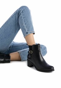 Botas de cuero negras hasta el tobillo con puntera redondeada, un tacón de bloque de altura media y un acento de hebilla dorada en el tobillo. Combinadas con unos vaqueros azules.