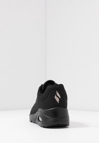 Scarpa sportiva nera con finitura liscia e opaca, forma arrotondata e un logo decorativo in oro rosa metallico sul tallone.
