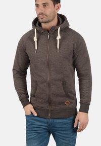 Bruine hoodie met een volledige rits, trekkoordaccenten, zijzakken en een geweven logo-embleem. Gladde textuur met een casual ontwerp.