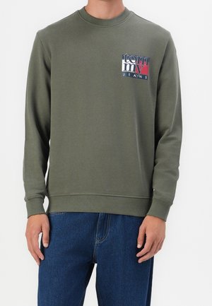 Mann trägt olivgrünes Sweatshirt mit Rundhalsausschnitt und "Tommy Jeans"-Logo auf der linken Brust sowie blaue Jeans, steht vor einfarbigem Hintergrund.
