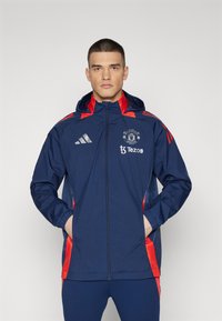 Chaqueta deportiva azul marino con detalles rojos, frente con cremallera y capucha. Presenta el logo del Manchester United y la marca Tezos en el pecho.