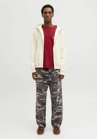 Haut de survêtement zippé de couleur claire, t-shirt bordeaux en dessous, et pantalon camouflé gris ample. Des chaussures en cuir marron à lacets complètent la tenue.