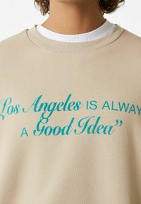 Felpa beige con testo turchese che recita "Los Angeles È SEMPRE UNA Buona Idea." Presenta un colletto rotondo e polsini a coste.