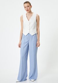 Gilet bianco in lino con scollo a V, caratterizzato da cinque bottoni. Abbinato a pantaloni a gamba larga azzurri e scarpe bianche a punta.