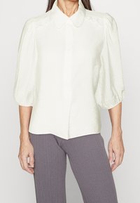 White puff-sleeve blouse with a button-up front, textured fabric, and a soft, rounded collar. Features delicate lace detailing along the shoulders.