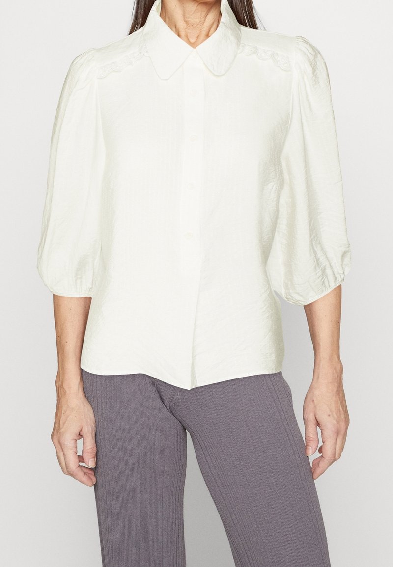 White puff-sleeve blouse with a button-up front, textured fabric, and a soft, rounded collar. Features delicate lace detailing along the shoulders.