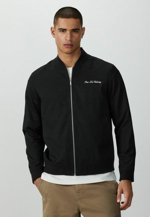 Junger Mann trägt eine schwarze Jacke mit Reißverschluss und weißen Schriftzügen auf der Brust, darunter ein weißes T-Shirt und Khakihosen, steht vor einem schlichten Hintergrund.