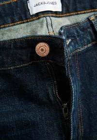 Jean en denim bleu foncé avec une fermeture éclair ouverte et un bouton en cuivre affichant la marque « JACK & JONES », et une étiquette en tissu blanc à l’intérieur de la ceinture.
