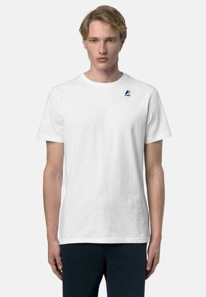 Giovane uomo che indossa una maglietta bianca semplice a maniche corte con un piccolo logo K-Way blu e rosso sul petto in alto a sinistra.