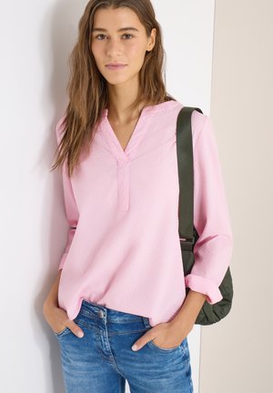 Ung kvinde med langt brunt hår iført en lyserød tekstureret bluse og blå jeans, lænende sig mod en hvid væg, med en grøn skuldertaske.