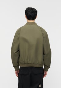 Άνδρας με στραμμένη πλάτη, φοράει μπουφάν τύπου bomber σε χακί χρώμα και μαύρο παντελόνι φορτίου, μπροστά σε απλό φωτεινό γκρι φόντο.