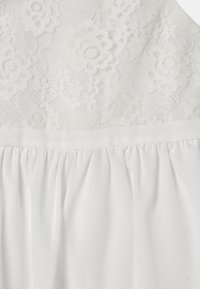 Vit klänning med en blommig spets-bodice med ett strukturerat mönster, kompletterad med en mjuk, rynkad kjol. Mjuk och lätt tyg.