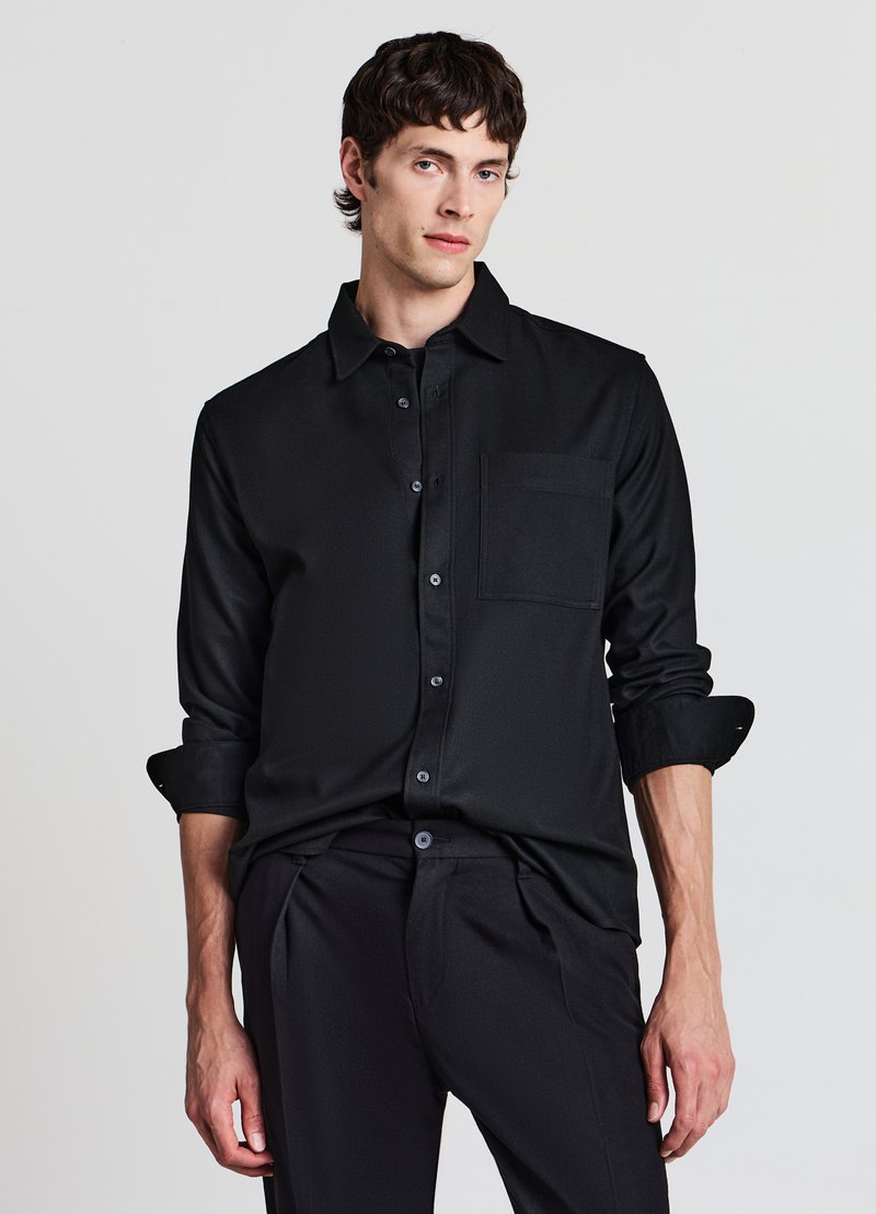 Schwarzes langärmeliges Hemd mit Knöpfen aus glattem Stoff, einer Brusttasche und hochgekrempelten Ärmeln, kombiniert mit passenden dunklen Hosen.