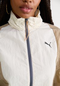 Lett, to-farget vindjakke med høy krage. Har full glidelås, oransje snor og en svart Puma-logo på brystet.