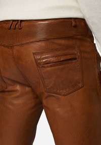 Bruine leren broeken hebben een gestructureerde tailleband en een paspelzak met een rits. Het gladde leer heeft een zachte glans en een minimale stiksels.