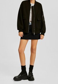 Schwarze Bomberjacke mit zwei Fronttaschen, gerippten Bündchen und einem durchgehenden Reißverschluss. Getragen über einem weißen T-Shirt, kombiniert mit einem schwarzen Minirock und klobigen Loafers.