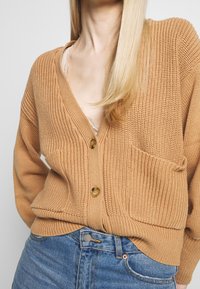 Locker sitzende Strickjacke in Hellbraun mit V-Ausschnitt, zwei Knöpfen und einer Fronttasche. Trägt man über einem Spitzenoberteil.