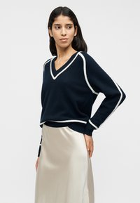 Πλεκτό μπλε σκούρο πουλόβερ με λαιμόκοψη σε σχήμα V και λευκή λεπτομέρεια, άνετη εφαρμογή, συνδυασμένο με μια λεία, ανοιχτόχρωμη σατέν φούστα.