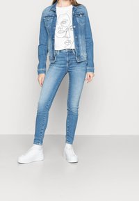 Vaaleansininen farkkutakki nappeineen, joka on puettu valkoisen t-paidan päälle, jossa on jatkuva viivapiirros kasvoista. Tiukat siniset farkut ja valkoiset lenkkarit.