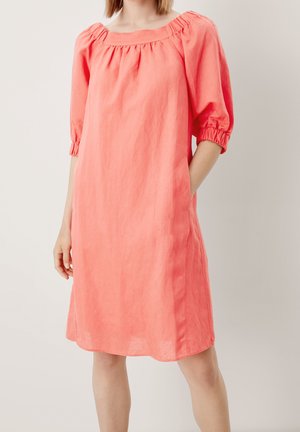 Žena v korálově zbarvených šatech délky po kolena s nařaseným výstřihem a tříčtvrtečními nabíranými rukávy, s rukou v kapse, stojící před jednobarevným pozadím.