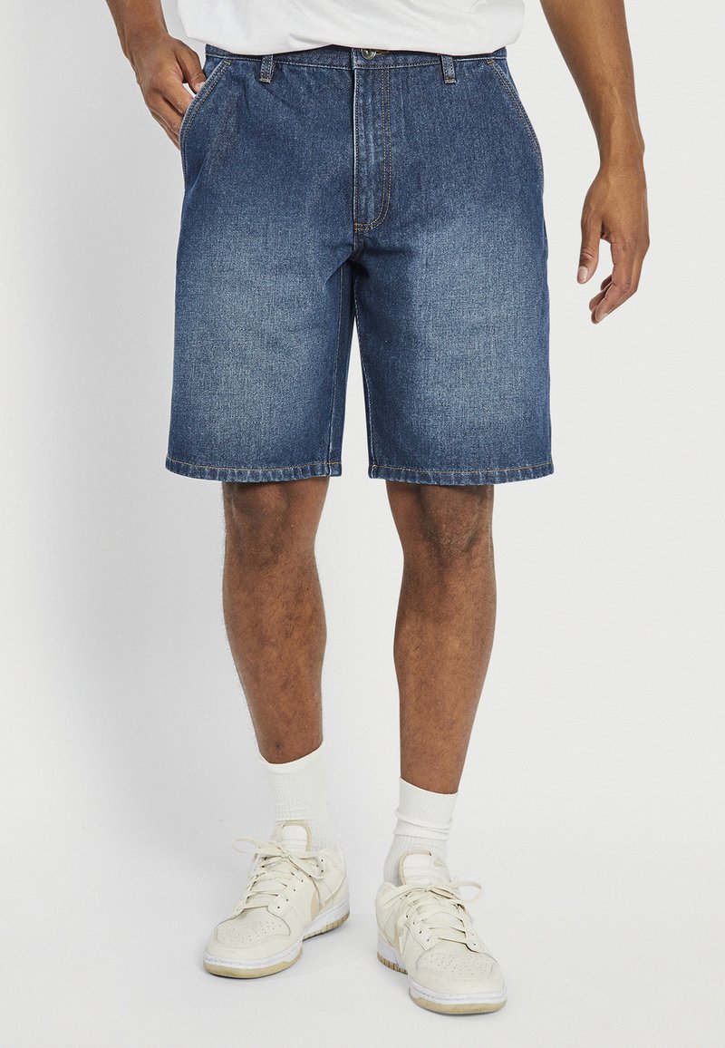 Denim shorts in dark blue, featuring a straight cut, five-pocket design, and stitched hem. Worn with white athletic socks and beige sneakers.