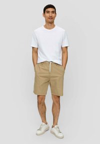 Weißes Baumwoll-T-Shirt kombiniert mit beigen Shorts, die einen elastischen Bund und einen Kordelzug haben. Weiße Sneakers runden das Outfit ab.