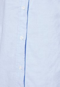 Chemise boutonnée bleu clair en tissu texturé. Comprend des boutons blancs, un col structuré et des coutures visibles le long de la patte de boutonnage.